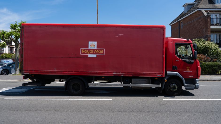 A red Royal Mail delivery truck parked on a residential street with houses and greenery visible in the background under a clear blue sky. The truck is positioned parallel to the road, with the side view fully visible, showing the Royal Mail logo and branding. The vehicle is on a paved road with marked lanes, and no pedestrians or other vehicles are in the immediate vicinity. The truck's cargo area is closed, indicating it is in transit or waiting to unload. This image depicts logistics and transportation related to home relocation or furniture transport services, as part of the moving process supported by Removal Companies Hammersmith.