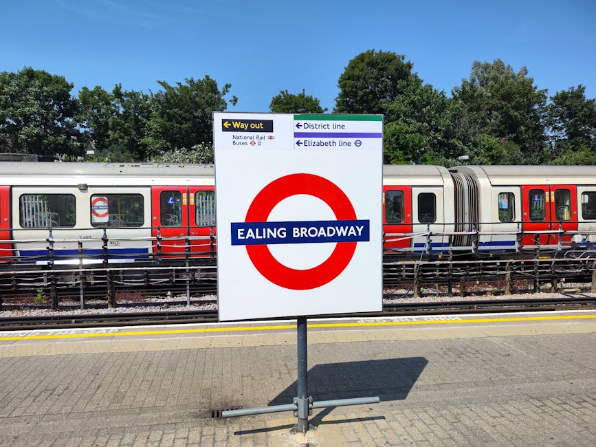 A large square railway station sign displaying 'Ealing Broadway' with a red London Underground roundel, situated on a paved platform with yellow safety lines. Behind the sign, a modern passenger train with silver bodies, red and blue accents, and multiple carriage doors is visible, parked on the tracks. The train features several windows, some of which are open, revealing the interior, and appears to be stationary. To the left of the sign, there is a small black and yellow sign indicating the way out, along with additional signage pointing to the district and Elizabeth lines. The background includes green, leafy trees under a clear blue sky, suggesting a bright, sunny day. The scene is associated with the London transportation hub, relevant to ongoing house removals or home relocation services nearby, as supported by the website removalcompanieshammersmith.co.uk.