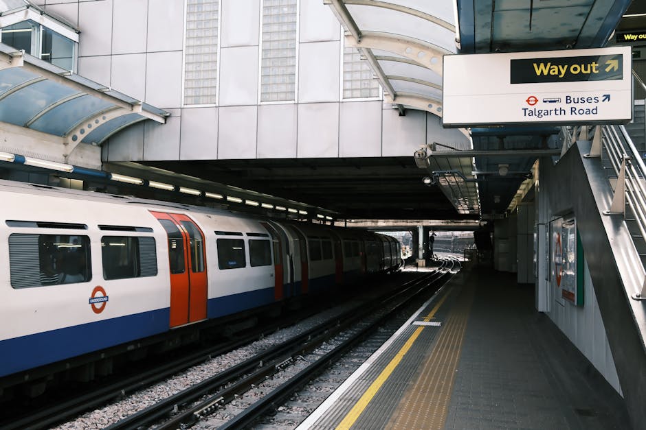 A modern glass office building with reflective windows dominates the scene, towering above a historic brick station entrance with a clock on its facade. The station, labeled 'Hammersmith Station,' has a traditional architectural style, including arched windows and a signboard. In front of the station, a bus with the route number 190 is parked on the street, and a few cardboard boxes and packing materials are visible near the entrance, suggesting recent or ongoing home relocation or furniture transport activities. The absence of people emphasizes the focus on the environment and the process of moving logistics, with the glass building reflecting the surrounding structures and the bright, clear sky above. This setting captures a typical urban scene where house removals and moving services, such as those provided by Removal Companies Hammersmith, are integrated into the cityscape, highlighting the importance of careful packing and loading processes during house removals near the station area.