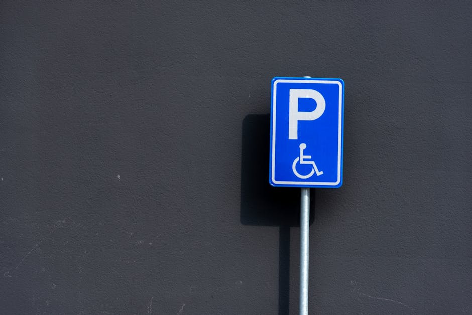 A blue parking sign with a white letter 'P' and a wheelchair symbol indicating accessible parking, mounted on a metal pole positioned against a smooth, dark grey wall. The sign is outdoors, with natur