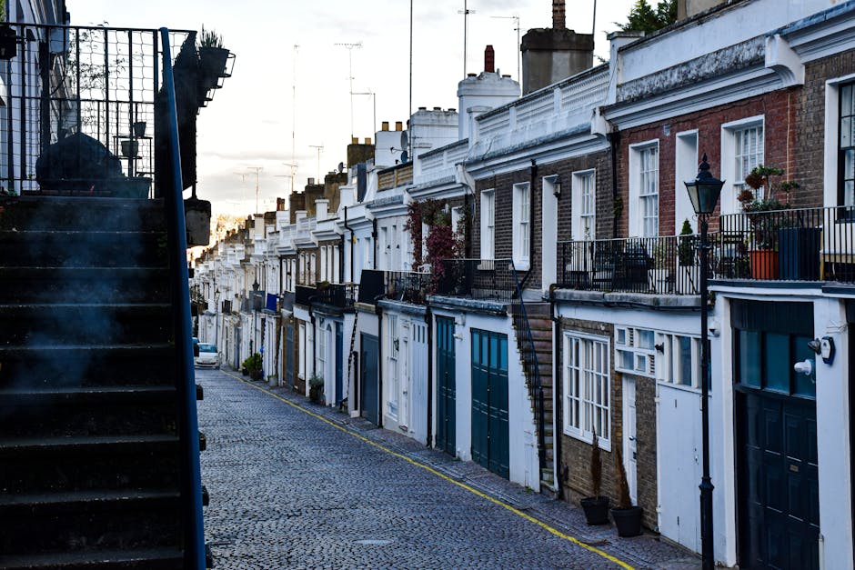 A narrow residential street in Hammersmith with a row of brick and stucco terraced houses featuring small front gardens, some with potted plants and outdoor lighting. The street has cobblestone paving and a slight incline, with a black metal staircase on the left side leading up to the first-floor entrances. One side of the street shows the front of houses with large windows, some with exterior lighting fixtures, and a mix of white and dark-colored doors. The rooftops are lined with chimneys and television antennas, and the sky is overcast, casting a soft, diffused light over the scene. The image captures an outdoor setting typical of home relocation environments, illustrating the challenges of moving furniture and boxes along narrow, uneven pathways, with no visible moving equipment or personnel present. This scene is relevant for furniture transport and packing during house removals in urban areas, as handled by Removal Companies Hammersmith.