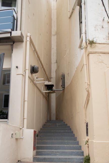A narrow outdoor staircase with metal steps ascending between two tall, beige-colored building walls. The left wall features visible white pipes, an electrical meter, and a small outdoor electrical box attached to the wall, while the right wall appears plain with some minor surface imperfections and a small plant growing near the base. The scene is illuminated by natural daylight, highlighting the confined space which presents challenges for furniture transport during home relocation. This image, related to King Street Hammersmith removal tips for narrow stairs, illustrates the logistical considerations involved in loading and moving large items through tight staircases. Removal Companies Hammersmith would typically manage such a loading process, ensuring proper handling and safety during house moves involving limited access.