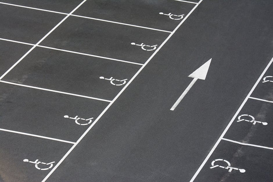 A section of a paved parking lot featuring several designated parking spaces marked with white lines and wheelchair symbols, indicating accessibility areas. In the center of the image, a white arrow painted on the asphalt points forward to guide traffic flow. The surface appears clean and well-maintained, with consistent marking paint. The surrounding area is part of an outdoor loading zone near a residential property, where Removal Companies Hammersmith is preparing for home relocation activities, including the loading of furniture and moving boxes into vans. This setting illustrates a common scene during the packing and moving process, emphasizing the importance of accessible parking for efficient house removals and furniture transport in the Hammersmith area, as detailed in the Ravenscourt Park removals parking and access advice.