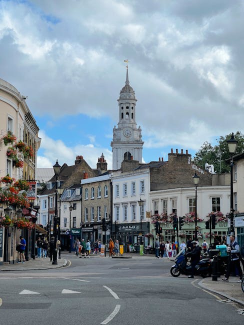 A busy street scene in Hammersmith during daytime, featuring a row of multi-storey buildings with retail shops on the ground floor and residential or office spaces above. The buildings are constructed from brick and light-colored materials, with some storefronts displaying signage and awnings. Pedestrians are walking along the sidewalk, some carrying bags or pushing strollers, while others are engaged in conversation. A few people are seen waiting at a crosswalk, where a black van and several motorcycles are parked at the curb. In the background, a prominent white clock tower with a gold weather vane at the top rises above the buildings, under a partly cloudy sky with patches of blue. Street lamps, flowering planters, and a bus stop sign line the street, indicating an active urban environment. The image highlights the typical setting for home relocation activities managed by Removal Companies Hammersmith, with furniture and packing materials potentially visible inside nearby properties, and the scene capturing the familiar city logistics involved in furniture transport and loading processes.