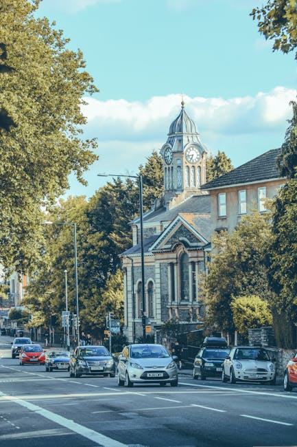 A busy street scene in Hammersmith during daytime, featuring a row of multi-storey buildings with retail shops on the ground floor and residential or office spaces above. The buildings are constructed from brick and light-colored materials, with some storefronts displaying signage and awnings. Pedestrians are walking along the sidewalk, some carrying bags or pushing strollers, while others are engaged in conversation. A few people are seen waiting at a crosswalk, where a black van and several motorcycles are parked at the curb. In the background, a prominent white clock tower with a gold weather vane at the top rises above the buildings, under a partly cloudy sky with patches of blue. Street lamps, flowering planters, and a bus stop sign line the street, indicating an active urban environment. The image highlights the typical setting for home relocation activities managed by Removal Companies Hammersmith, with furniture and packing materials potentially visible inside nearby properties, and the scene capturing the familiar city logistics involved in furniture transport and loading processes.