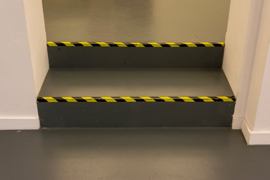 A close-up view of a set of indoor stairs with two steps, painted in dark grey, situated inside a property. The stairs are bordered by white walls on both sides and marked with yellow and black hazard tape along the edges to indicate caution during the moving process. The flooring at the base of the stairs is a smooth, light grey surface, likely concrete or vinyl. This image represents a typical challenge in house removals when navigating narrow or staircase access points during home relocation. The setting is well-lit, with no visible furniture or moving equipment present, focusing solely on the stairs and safety markings. This visual detail aligns with services offered by Removal Companies Hammersmith, highlighting the importance of careful logistics when handling furniture transport through tight or difficult access areas during relocations.