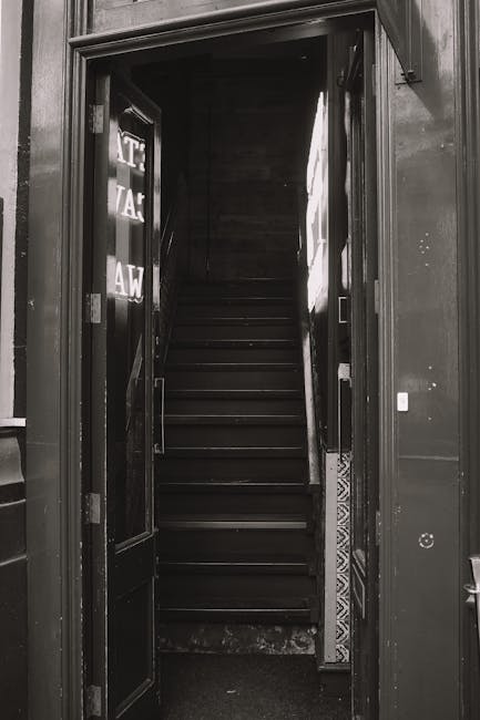 A narrow staircase inside a building, viewed from the ground floor doorway, with dark wooden steps ascending out of view. The staircase is enclosed by dark walls and a wooden railing on the right side. The entrance doorway is framed by a dark doorframe and features partially visible signage on the left door panel, which is open. The surrounding interior appears to be dimly lit, consistent with an indoor environment prepared for a home relocation or furniture transport. Removal Companies Hammersmith are likely managing the logistics of moving large items through this restricted access area, using equipment like trolleys or straps to facilitate the safe handling of furniture and boxes. The focus is on the tight, challenging passage typical of house removals involving staircase and narrow access, highlighting the importance of careful planning and professional packing and moving services for successful furniture transport.