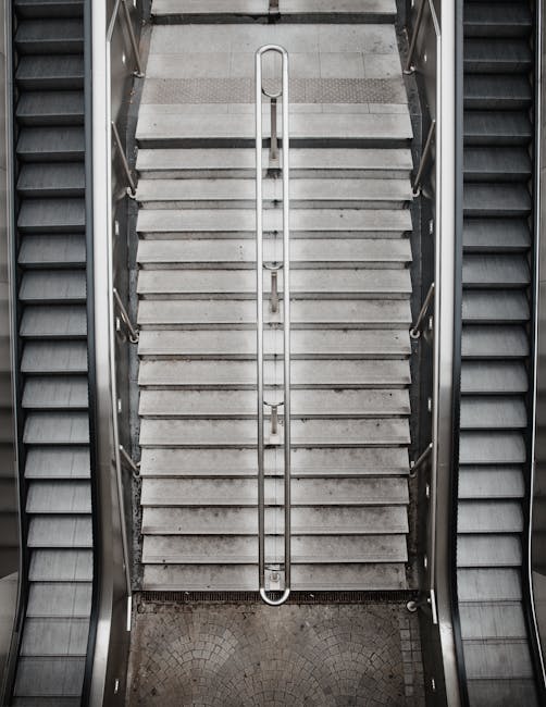 A close-up view of a set of indoor stairs with two steps, painted in dark grey, situated inside a property. The stairs are bordered by white walls on both sides and marked with yellow and black hazard tape along the edges to indicate caution during the moving process. The flooring at the base of the stairs is a smooth, light grey surface, likely concrete or vinyl. This image represents a typical challenge in house removals when navigating narrow or staircase access points during home relocation. The setting is well-lit, with no visible furniture or moving equipment present, focusing solely on the stairs and safety markings. This visual detail aligns with services offered by Removal Companies Hammersmith, highlighting the importance of careful logistics when handling furniture transport through tight or difficult access areas during relocations.