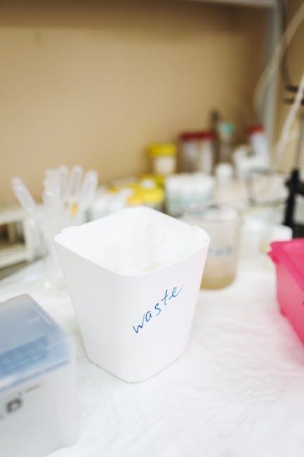 Inside a property, a white waste bin with the word 'waste' written in blue ink is positioned on a table covered with a white cloth. In the background, there are various containers and bottles with lid