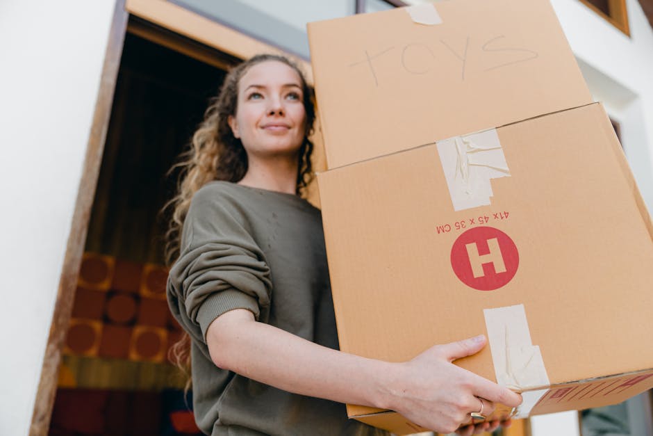 A young woman with long, curly brown hair is inside a home, holding a cardboard box labeled 'toys' secured with packing tape. She is wearing a grey sweatshirt with the sleeves rolled up and has a slight smile on her face. In the background, a doorway reveals a room with wooden paneling or furniture, and the lighting suggests natural daylight. The box is part of a moving process, likely during a home relocation or furniture transport, and there are additional cardboard boxes behind her, indicating packing and moving preparations. The setting is indoors, near an open entrance or hallway, and the overall scene captures an organized packing or loading process as part of house removals. This description aligns with services offered by Removal Companies Hammersmith, focusing on furniture transport and packing for relocations.